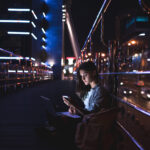 side view of young woman with laptop on knees using smartphone with night city on background
